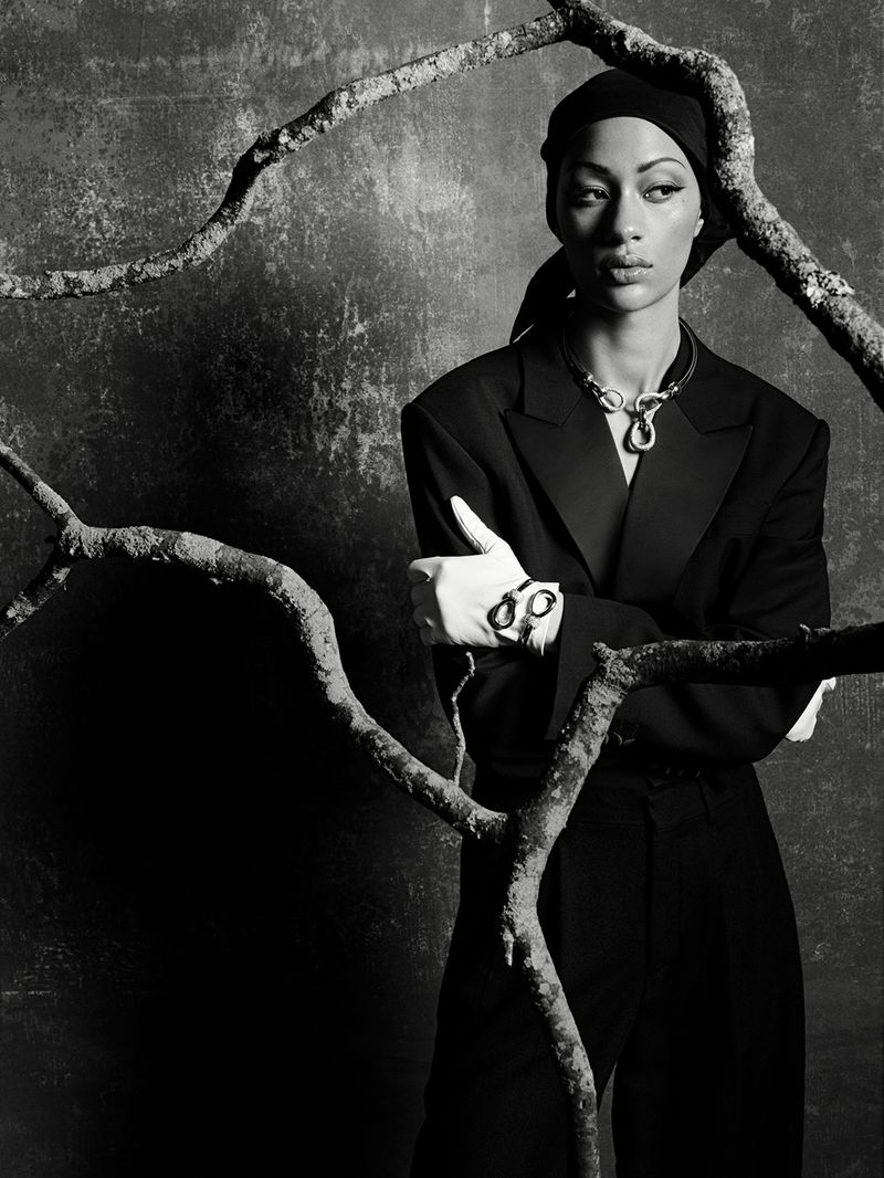 Model Selena Forrest in a black suit, headscarf, and white gloves framed by gnarly tree branches in a studio shoot.