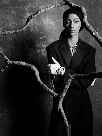Model Selena Forrest in a black suit, headscarf, and white gloves framed by gnarly tree branches in a studio shoot.