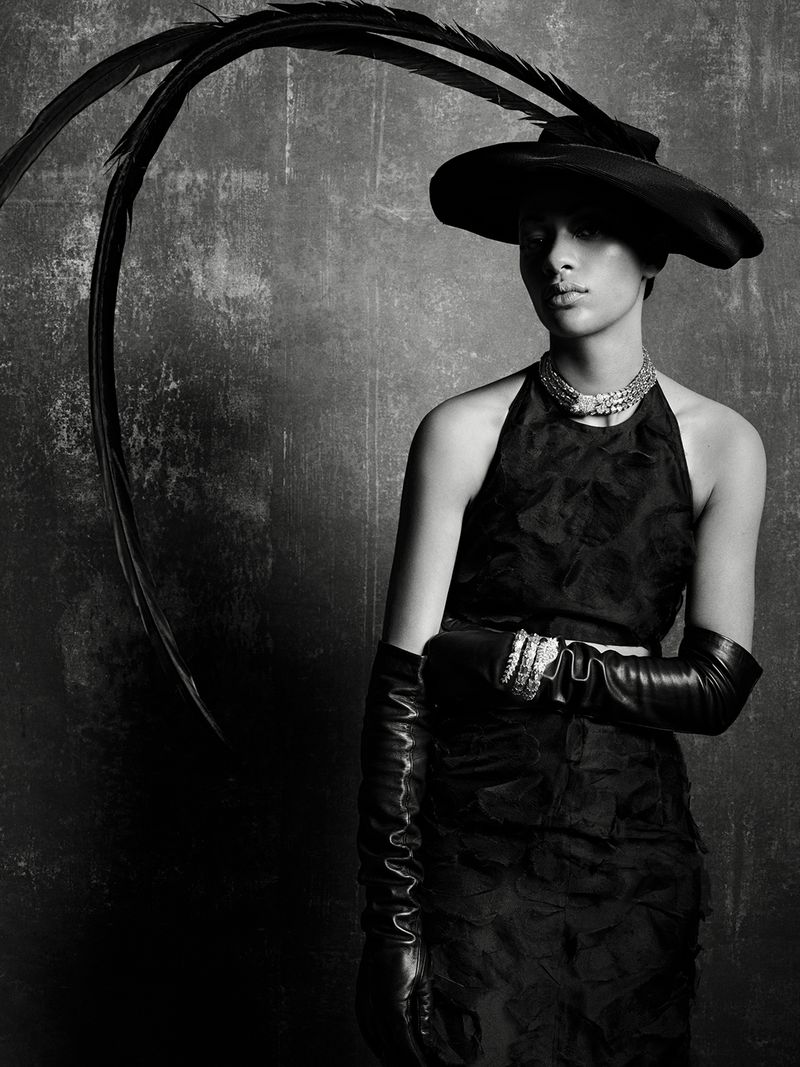 Model Selena Forrest wearing a black hat with long feathers, halter dress, and leather gloves against a textured background.