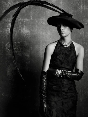 Model Selena Forrest wearing a black hat with long feathers, halter dress, and leather gloves against a textured background.