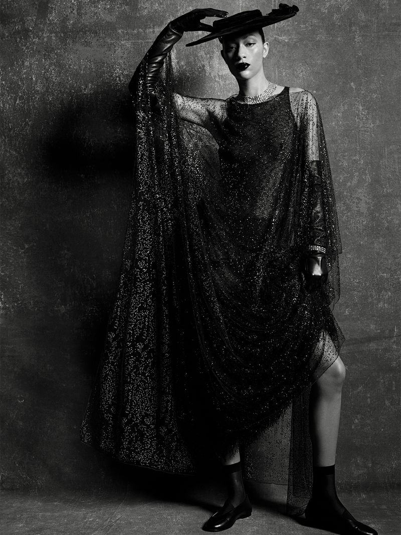 Model Selena Forrest wearing a black sheer glitter gown, wide-brim hat, and leather gloves against a dark textured background.