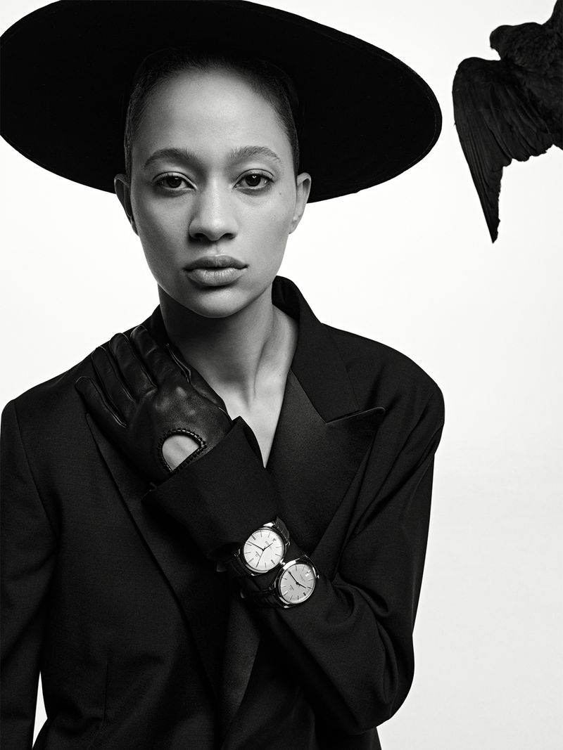 Model Selena Forrest wearing a wide-brimmed black hat, tailored jacket, leather glove, and watches against a white background.