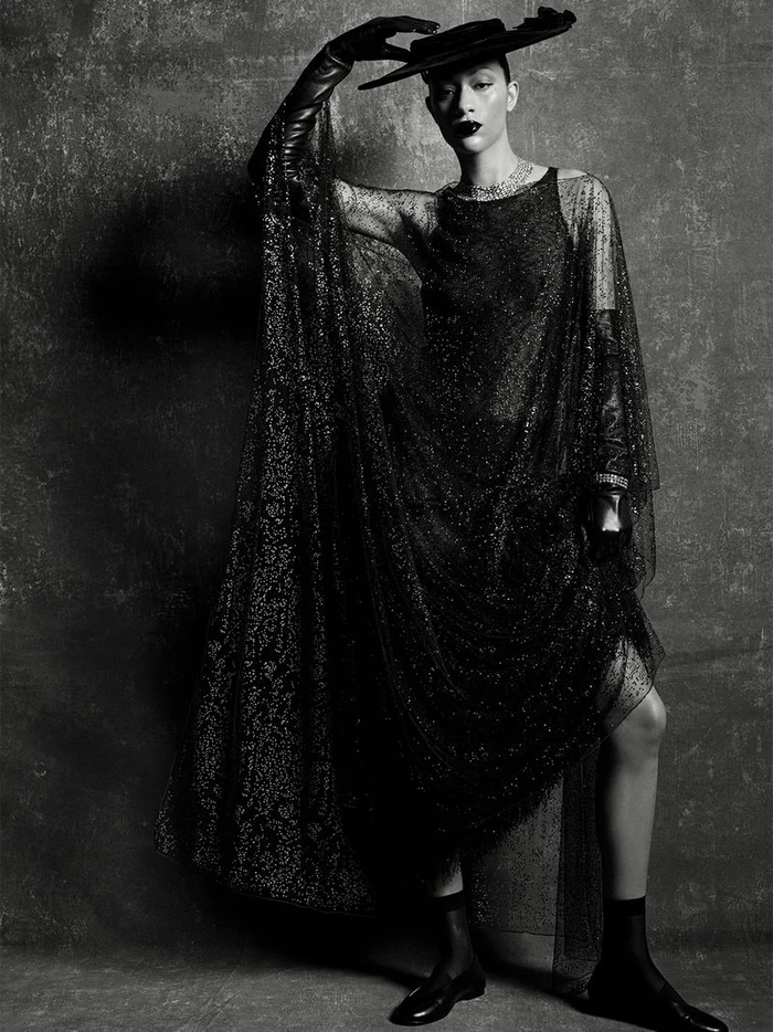 Model Selena Forrest wearing a black sheer glitter gown, wide-brim hat, and leather gloves against a dark textured background.