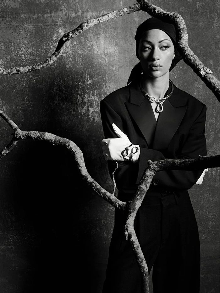 Model Selena Forrest in a black suit, headscarf, and white gloves framed by gnarly tree branches in a studio shoot.