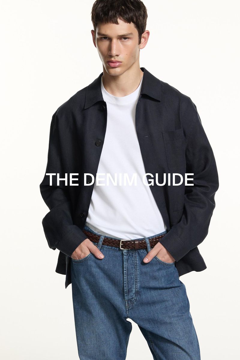 A young adult male poses in a casual fashion ensemble: light blue jeans, white t-shirt, and navy overshirt against a white backdrop.