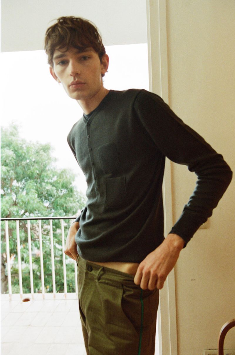 Young male model wearing a dark long-sleeve top and olive trousers in a light-filled interior with a balcony background