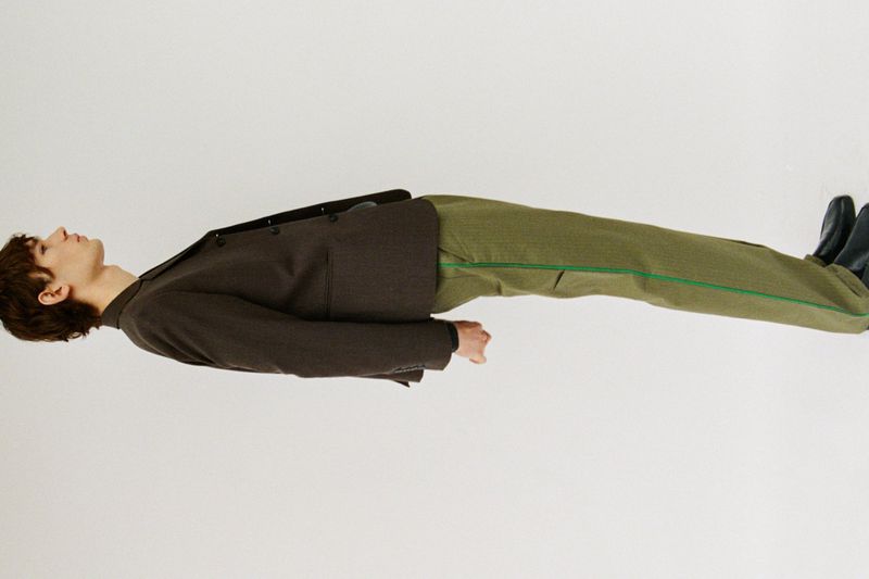 Model wearing a dark brown blazer and green trousers lying sideways against a seamless white background.