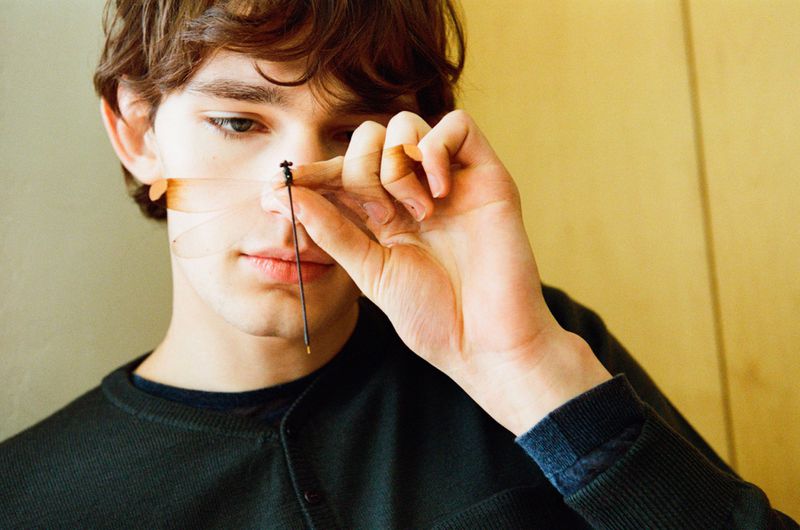 Close-up portrait of a young man wearing a dark sweater holding a delicate insect near his face for Paloma Wool Man Spring 2026.