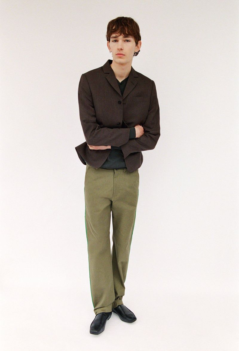 Model wearing a dark brown blazer and green trousers standing against a plain white background for Paloma Wool.