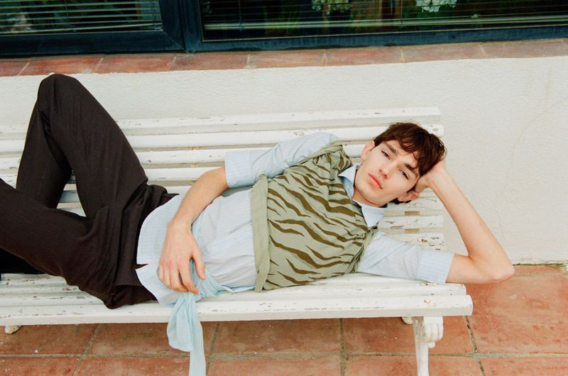 A young male model wearing patterned knit vest and dark trousers reclining on a white wooden bench outdoors.