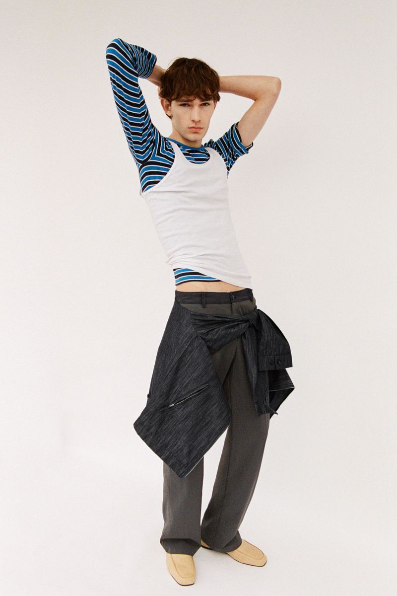 Male model with hands behind head wearing striped long sleeve top, white tank top, grey trousers tied with jacket at waist.