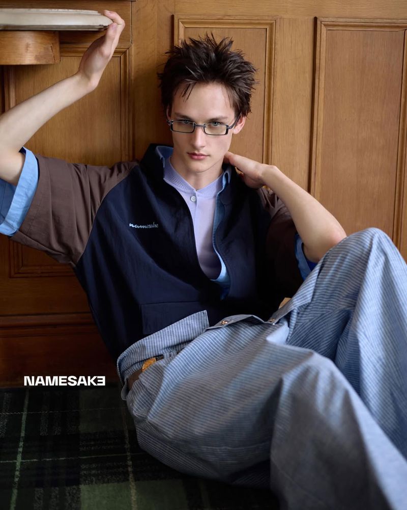 Young male model wearing glasses, layered polo shirt, and striped trousers seated against a wood-paneled wall