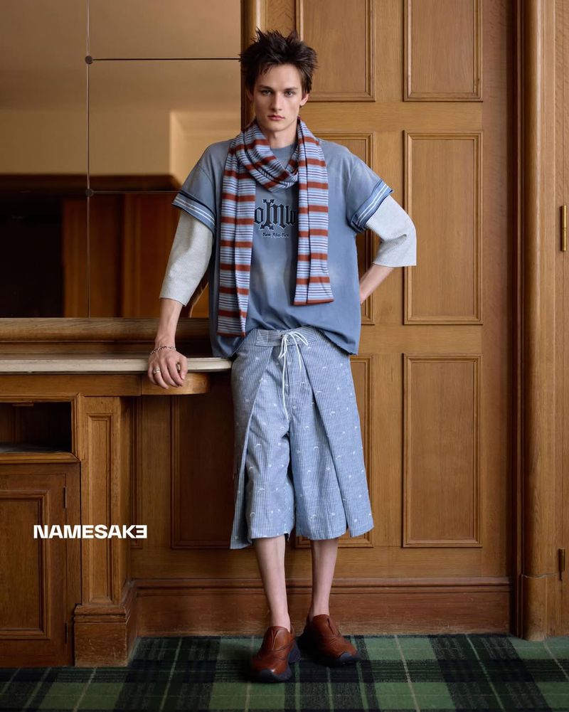 Male model wearing a blue layered t-shirt, striped scarf, and shorts standing indoors against wood paneling