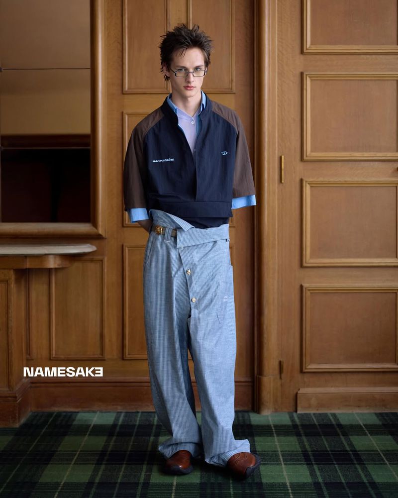 Young man in a blue, brown, and light blue short-sleeved jacket and light-blue baggy trousers stands in front of a wood paneled wall.