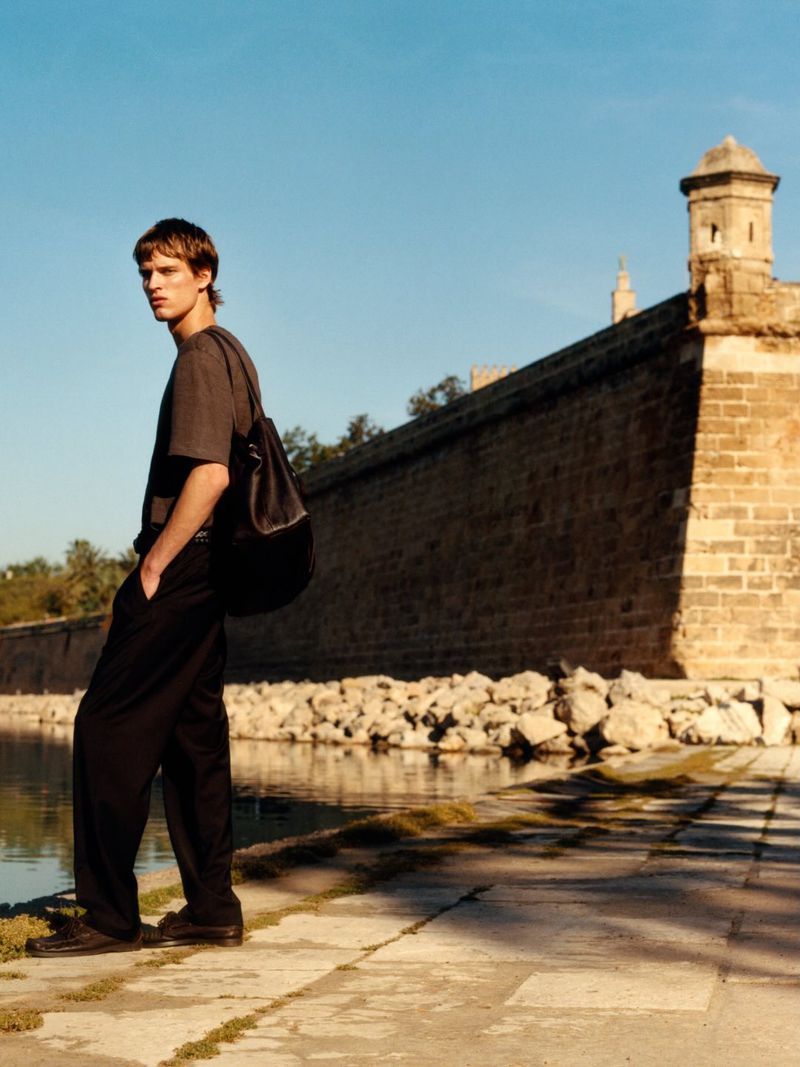 A male model standing by a stone fortress wall and water, wearing a dark grey t-shirt and black trousers with a leather shoulder bag.