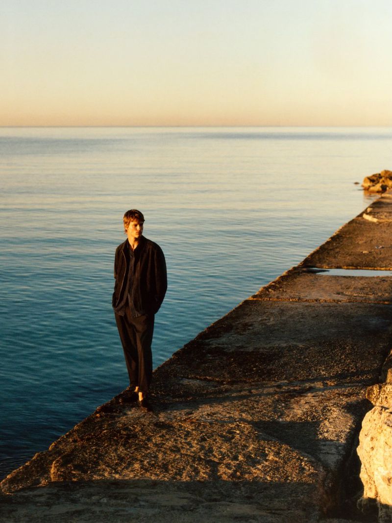 A young male model standing on a concrete pier overlooking calm ocean water under a sunset sky for Massimo Dutti