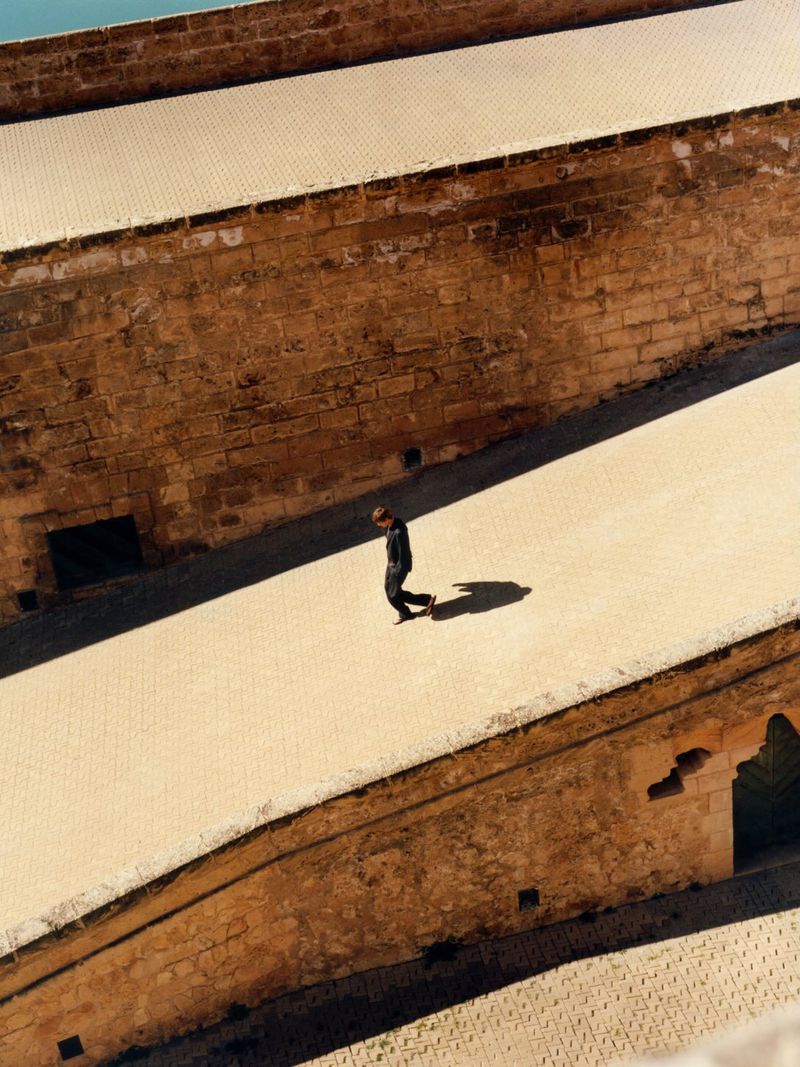 High-angle shot of a person walking along a stone pathway between large weathered walls for Massimo Dutti Mallorca Outline S/S 26