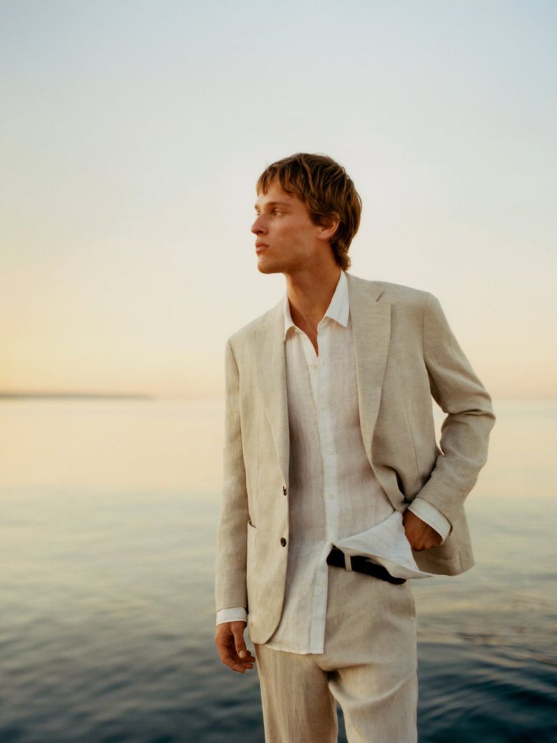 A male model wearing a beige linen suit and white shirt standing against an ocean and sunset sky background.