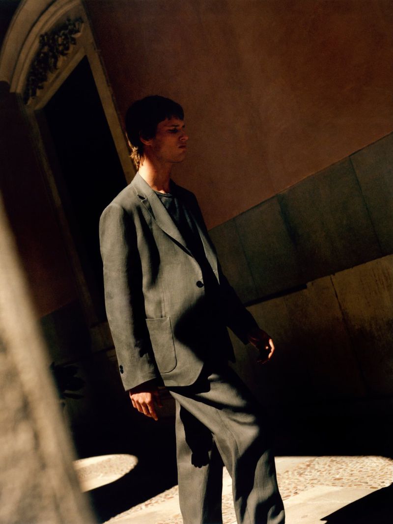 Man wearing a grey suit walking outdoors under dramatic sunlight with warm stone architecture in the background.