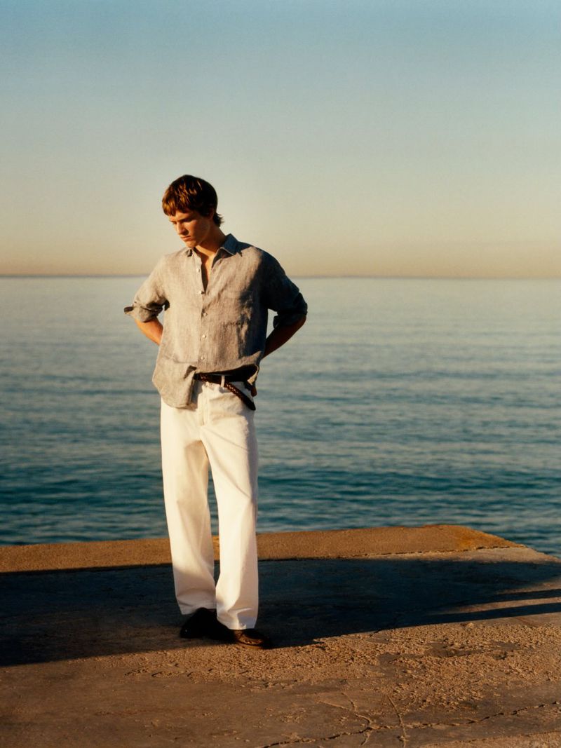 Model wearing a grey button-down shirt and white trousers standing by the sea during sunset for Massimo Dutti