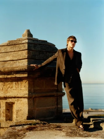 An adult male in a dark suit poses near a stone fortress overlooking the ocean on a sunny day, showcasing a blend of fashion and architecture.