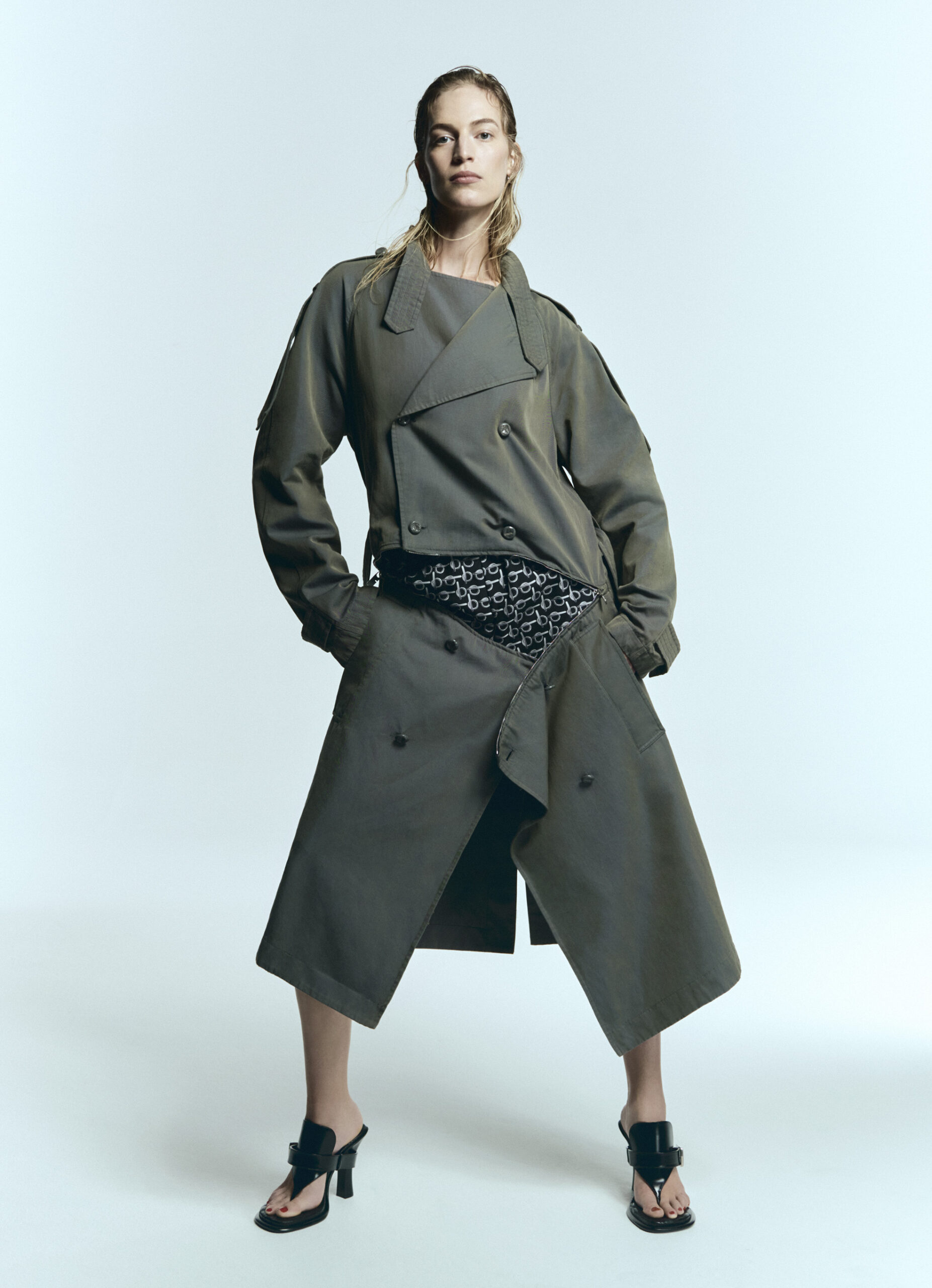 Fashion shot of a blonde adult woman wearing a trench coat and open-toe heeled sandals, posing against a white backdrop in a studio.