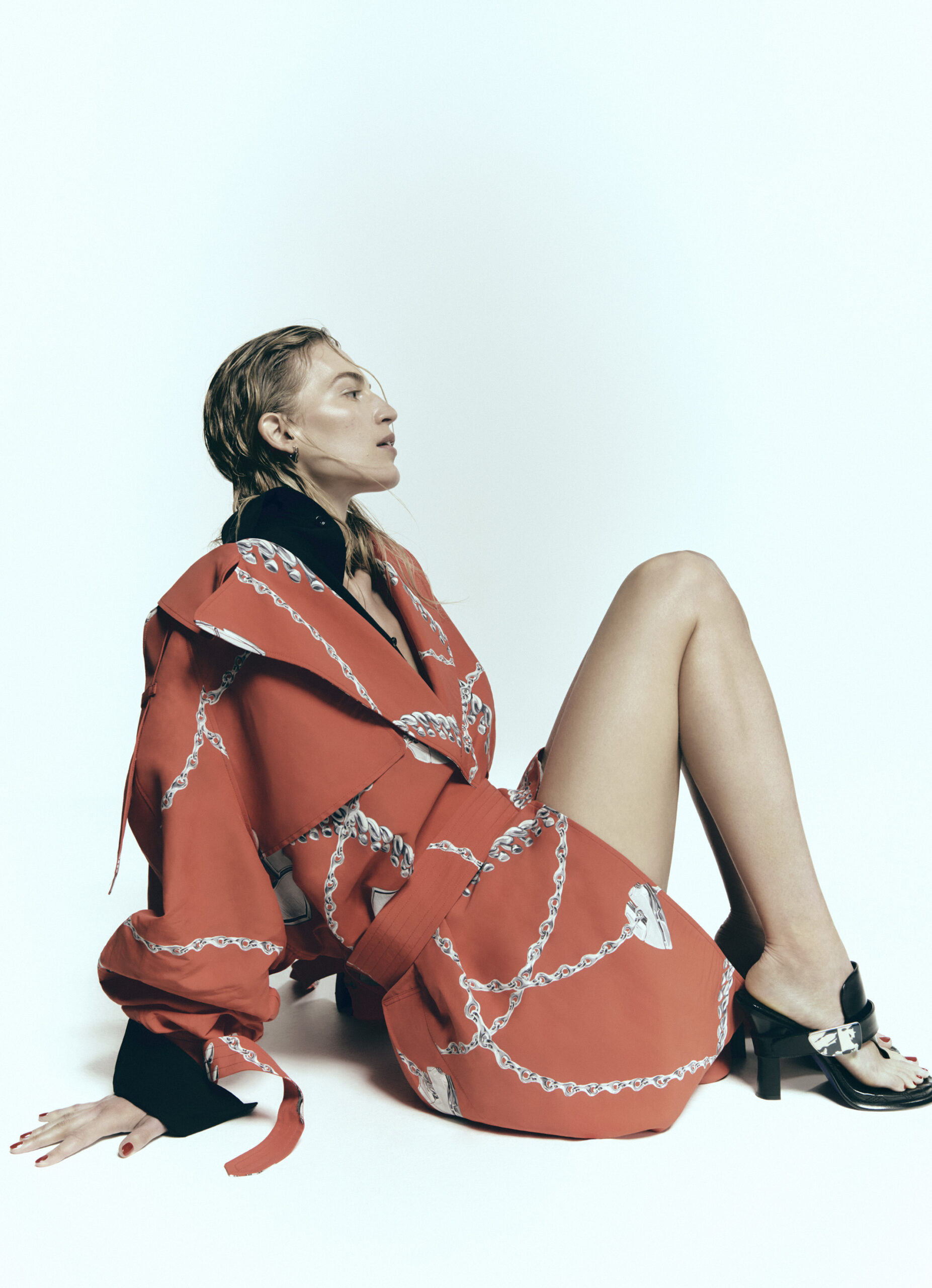 A woman sits against a white background wearing a chain-patterned jacket and skirt, complemented by black heeled sandals and wet styled hair.
