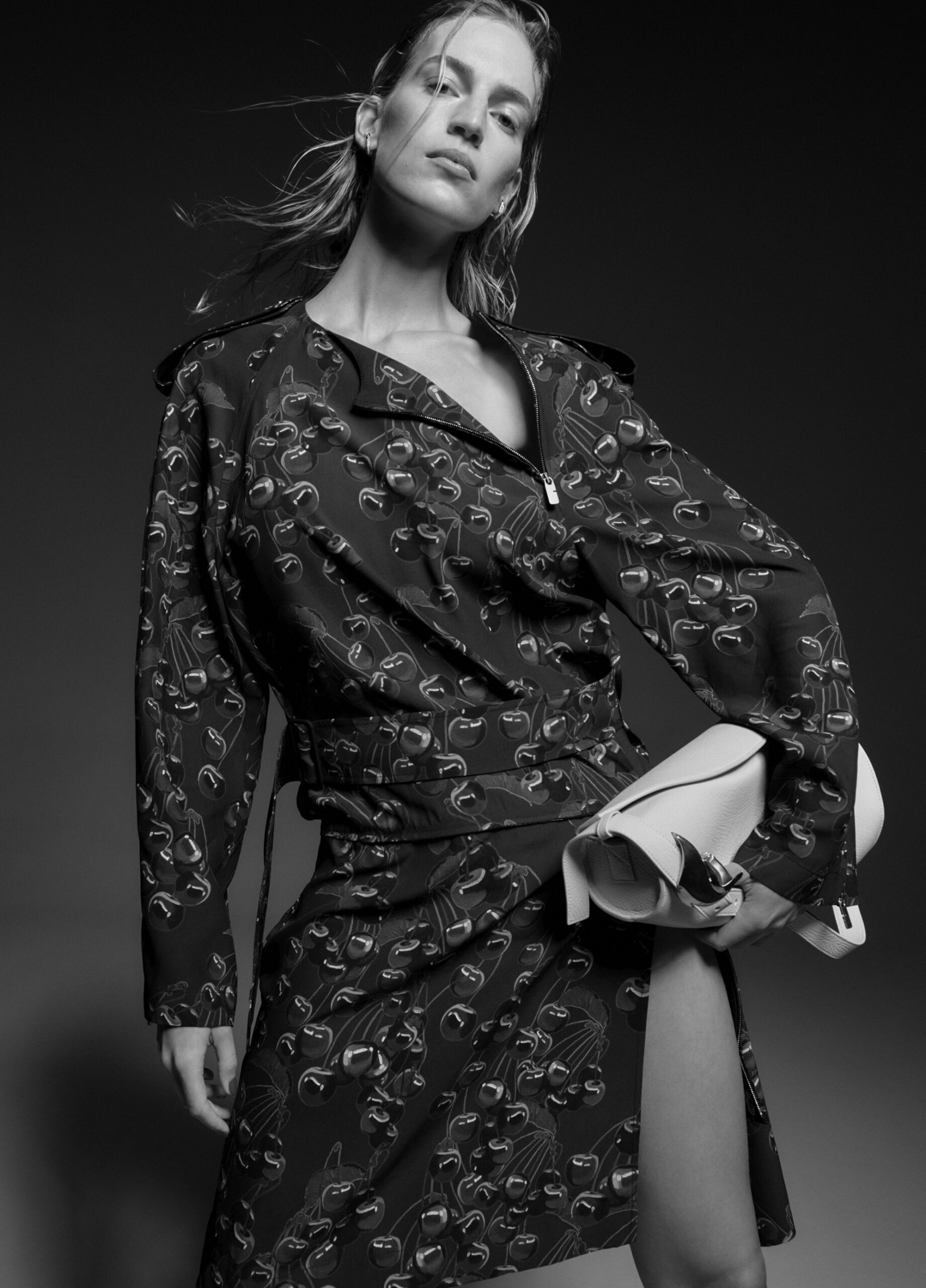 Monochrome studio portrait of a blonde adult female fashion model wearing a patterned wrap dress and holding a light-colored clutch purse.