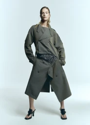 Model wearing an olive green belted trench coat and black heeled mules against a white seamless background
