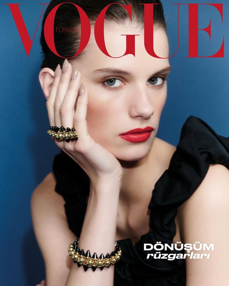 Close-up portrait of a model with red lipstick, holding her hand to her cheek, wearing a black top and spiked bracelets, against a blue background