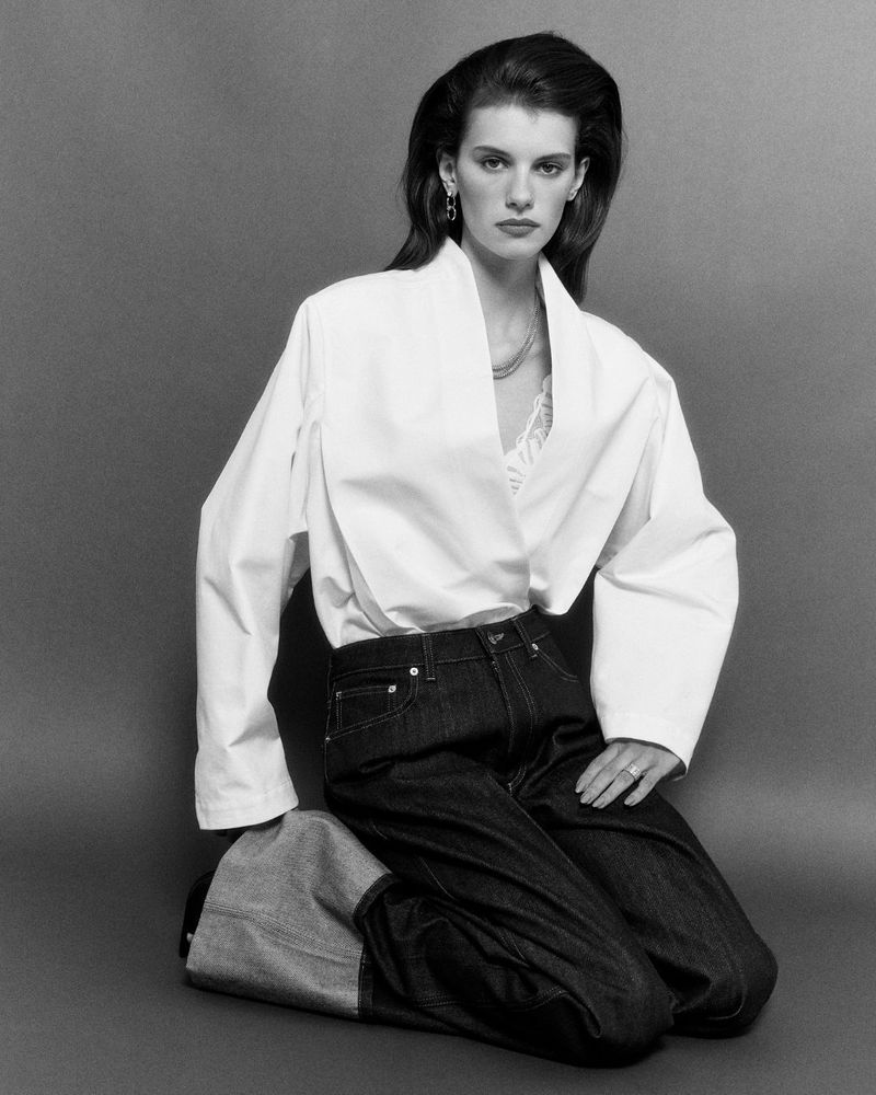 Model kneeling in white structured blouse and dark trousers against a plain grey studio backdrop