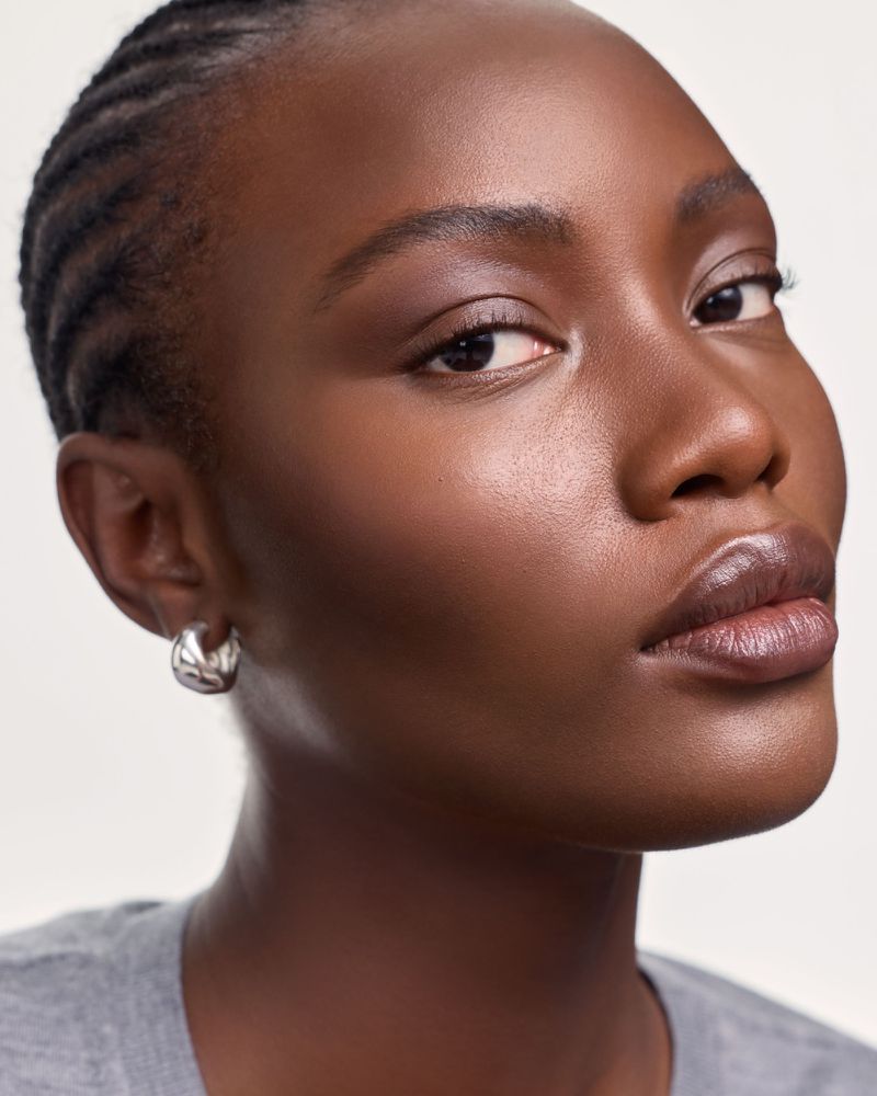 Close-up portrait of a Black woman with braided hair, wearing silver hoop earrings and a gray shirt against a neutral background.
