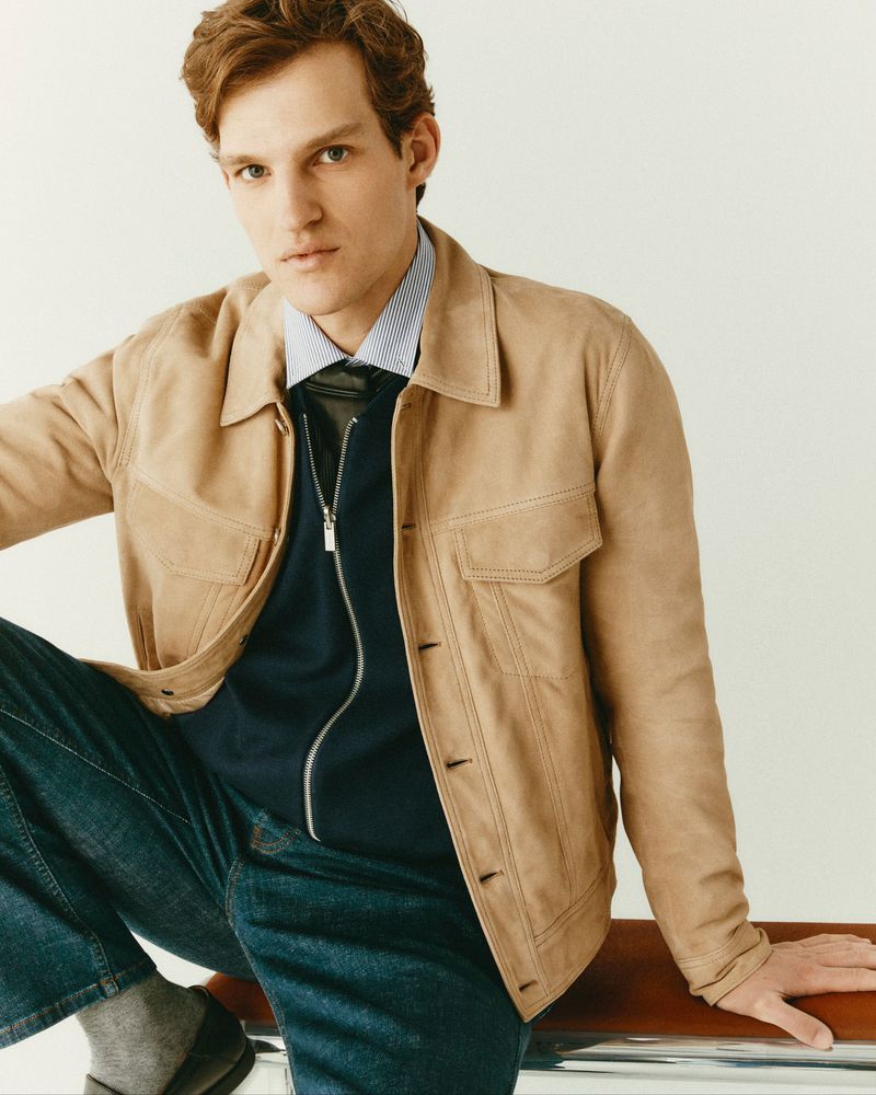 Man wearing camel suede jacket, striped shirt, and blue jeans seated indoors against a plain light wall.