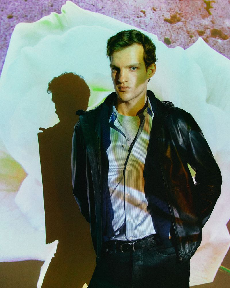 Studio portrait of a handsome man in a striped shirt and leather jacket, posing in front of a flower pattern wall.