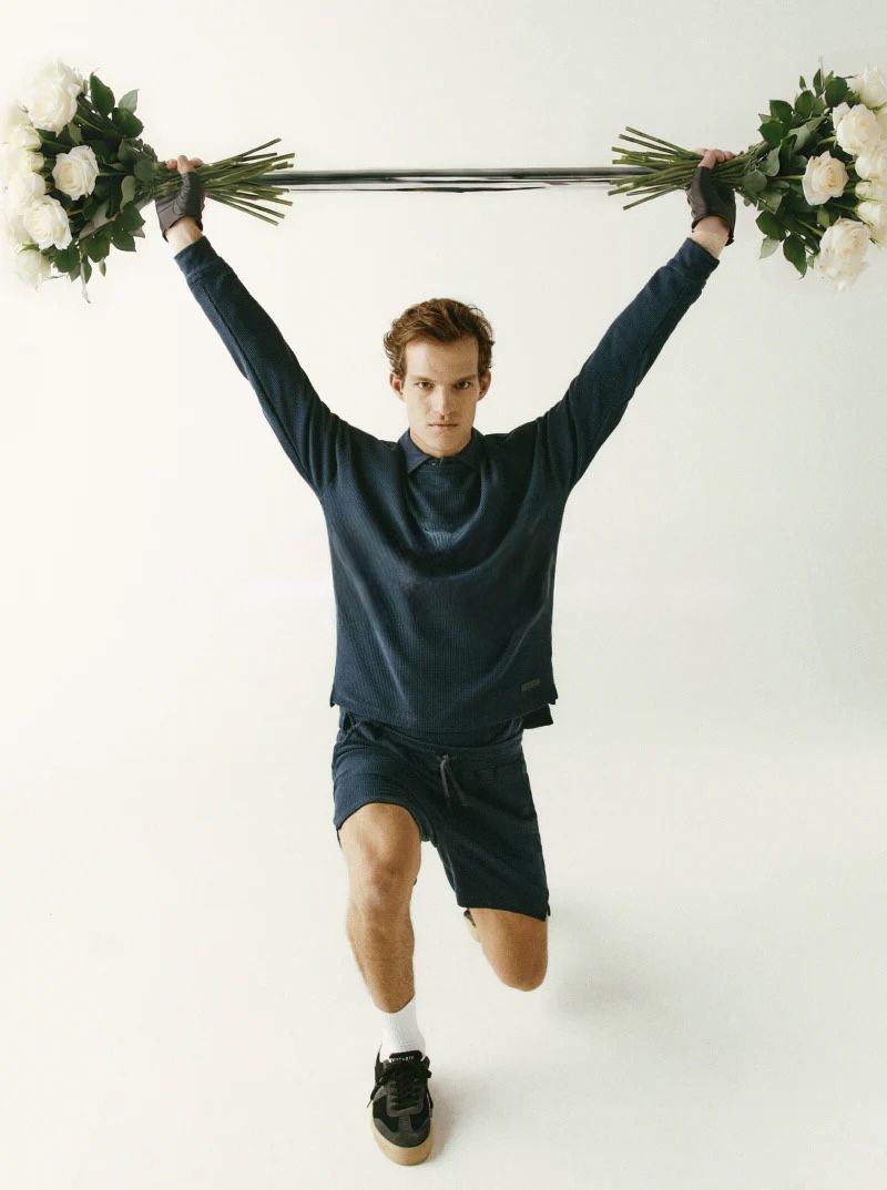 Model lifting a barbell loaded with large white rose bouquets on a white studio background for Trussardi.