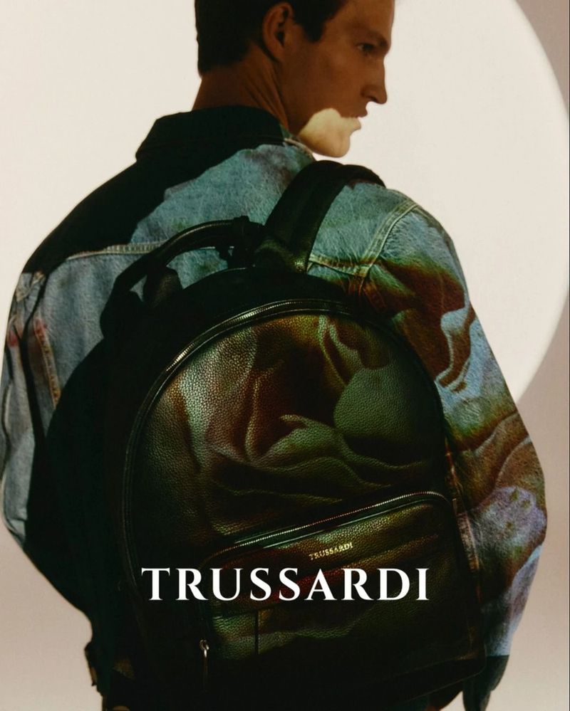 Model wearing a printed denim jacket carrying a large black leather backpack with Trussardi branding against a light background