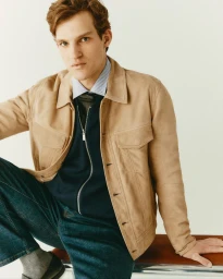 Man wearing camel suede jacket, striped shirt, and blue jeans seated indoors against a plain light wall.