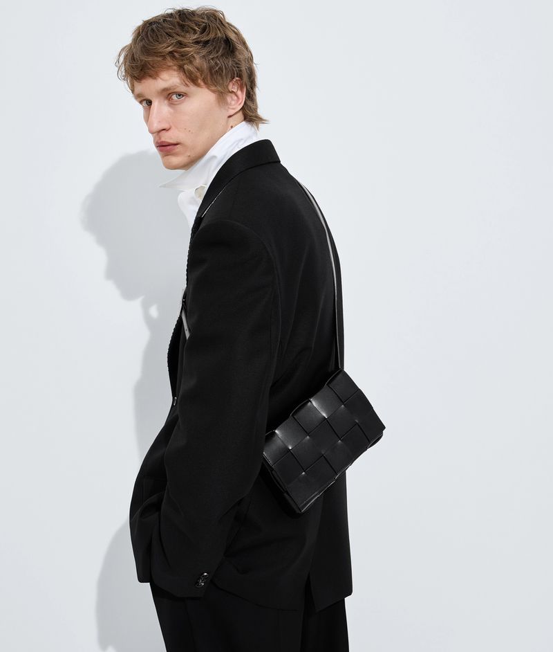 Male model wearing black blazer and white shirt with woven black leather crossbody bag against white background