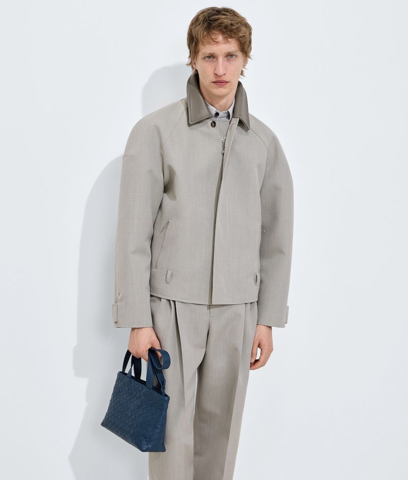 Model wearing a grey jacket and trousers holding a blue handbag against a white studio background.