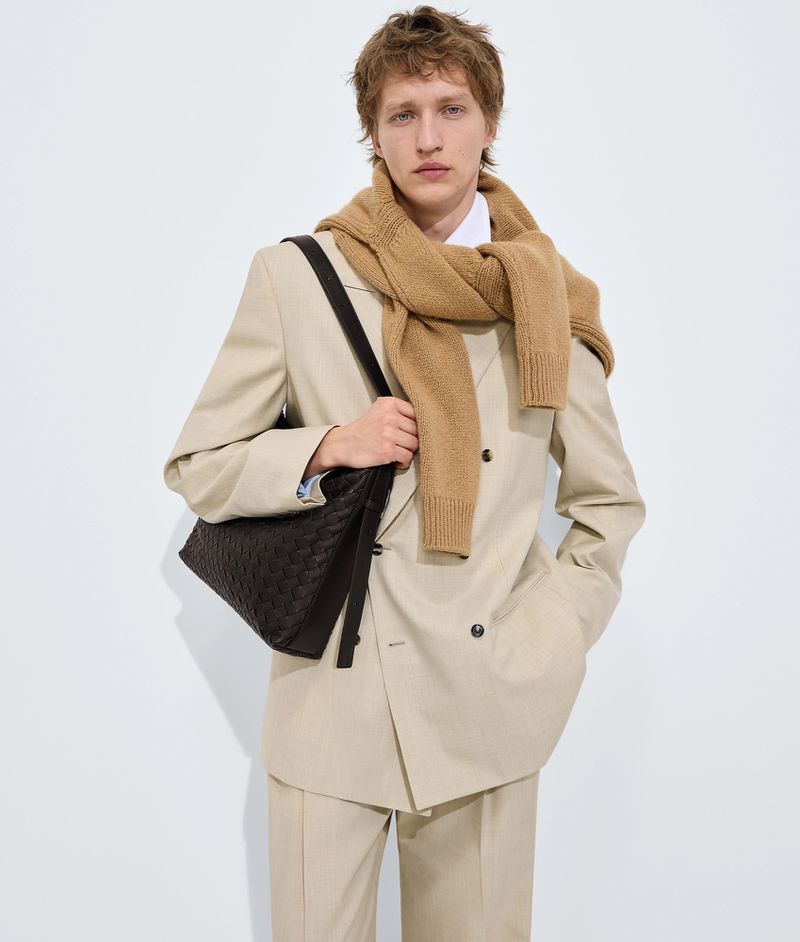 Model wearing a light beige suit and camel sweater with a dark woven leather shoulder bag against a white seamless background
