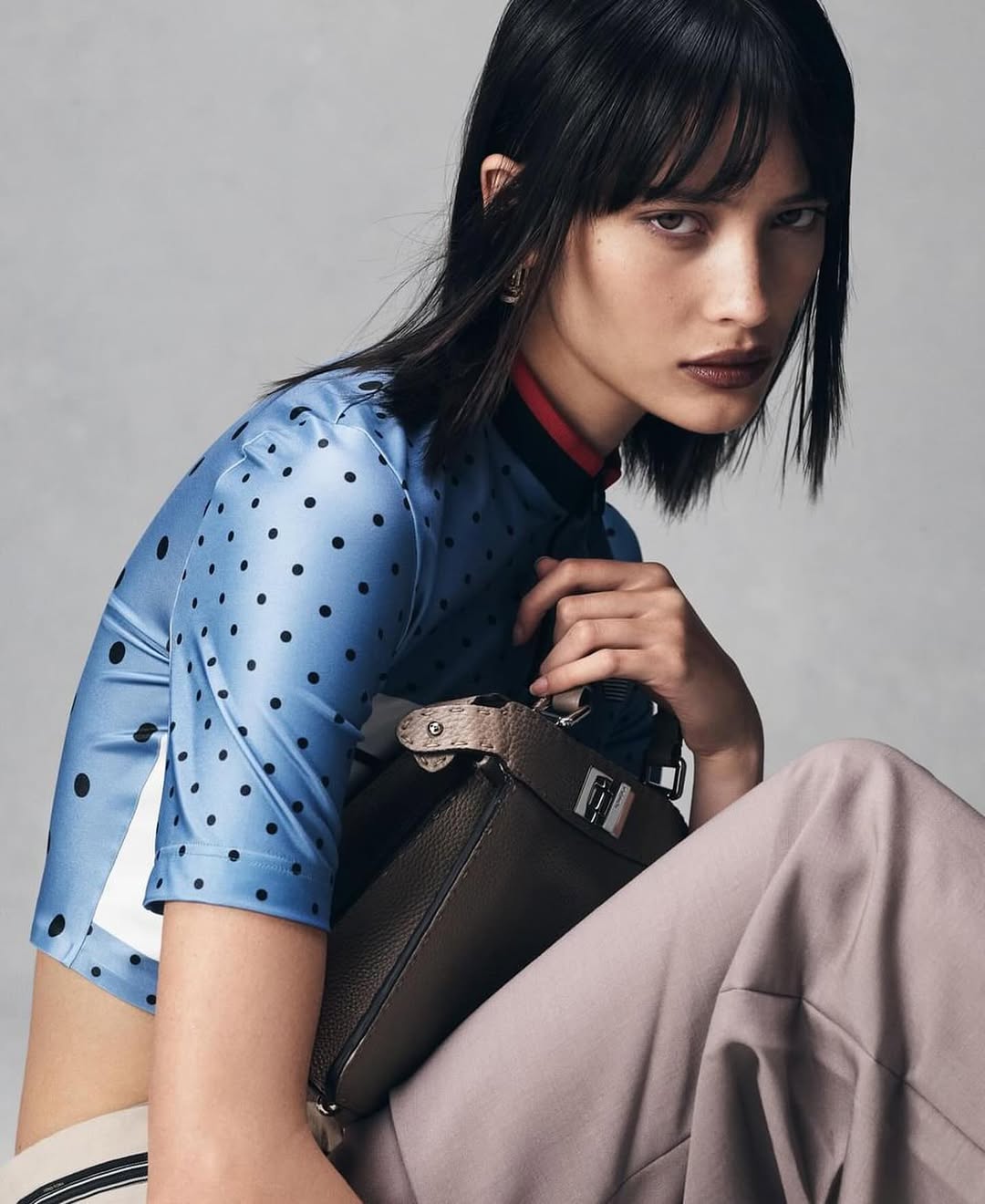 A woman in a blue polka-dot top poses with a brown handbag in a studio shot. Her gaze is directed towards the camera, and she wears stylish makeup.