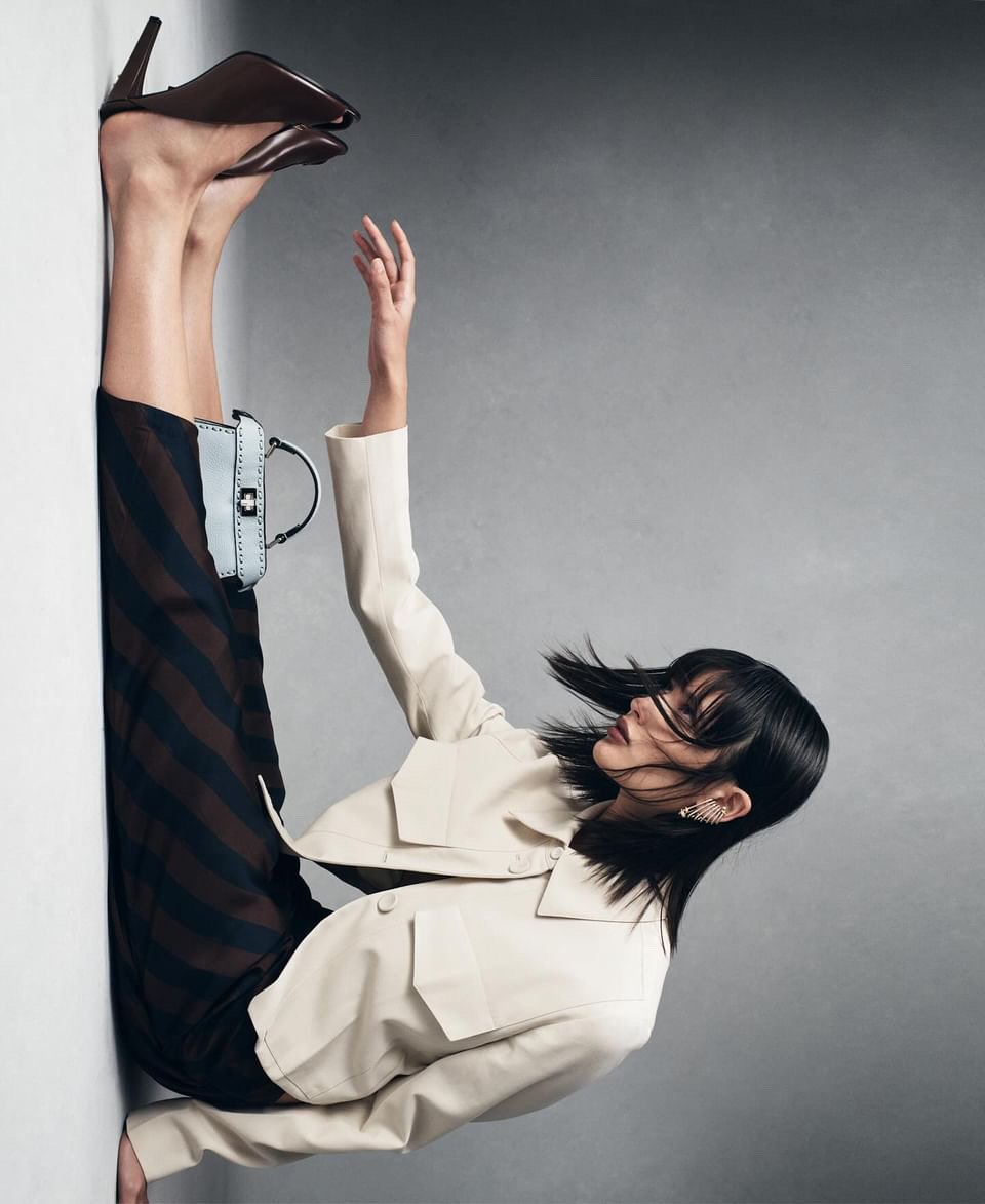 Fashionable woman strikes a pose in a studio. She wears a cream jacket, striped skirt, and brown heels, while a light blue purse adorns the scene.