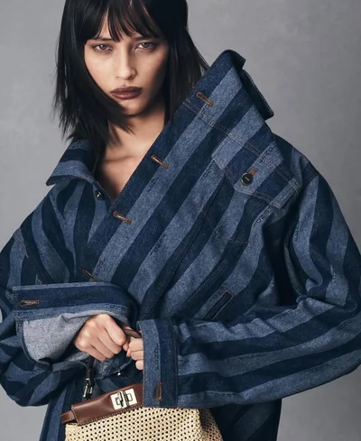 Model in striped denim jacket holding a structured handbag against a grey textured studio backdrop