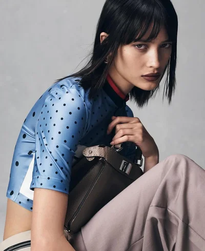 Model in a blue polka dot top holding a dark handbag against a neutral grey studio background