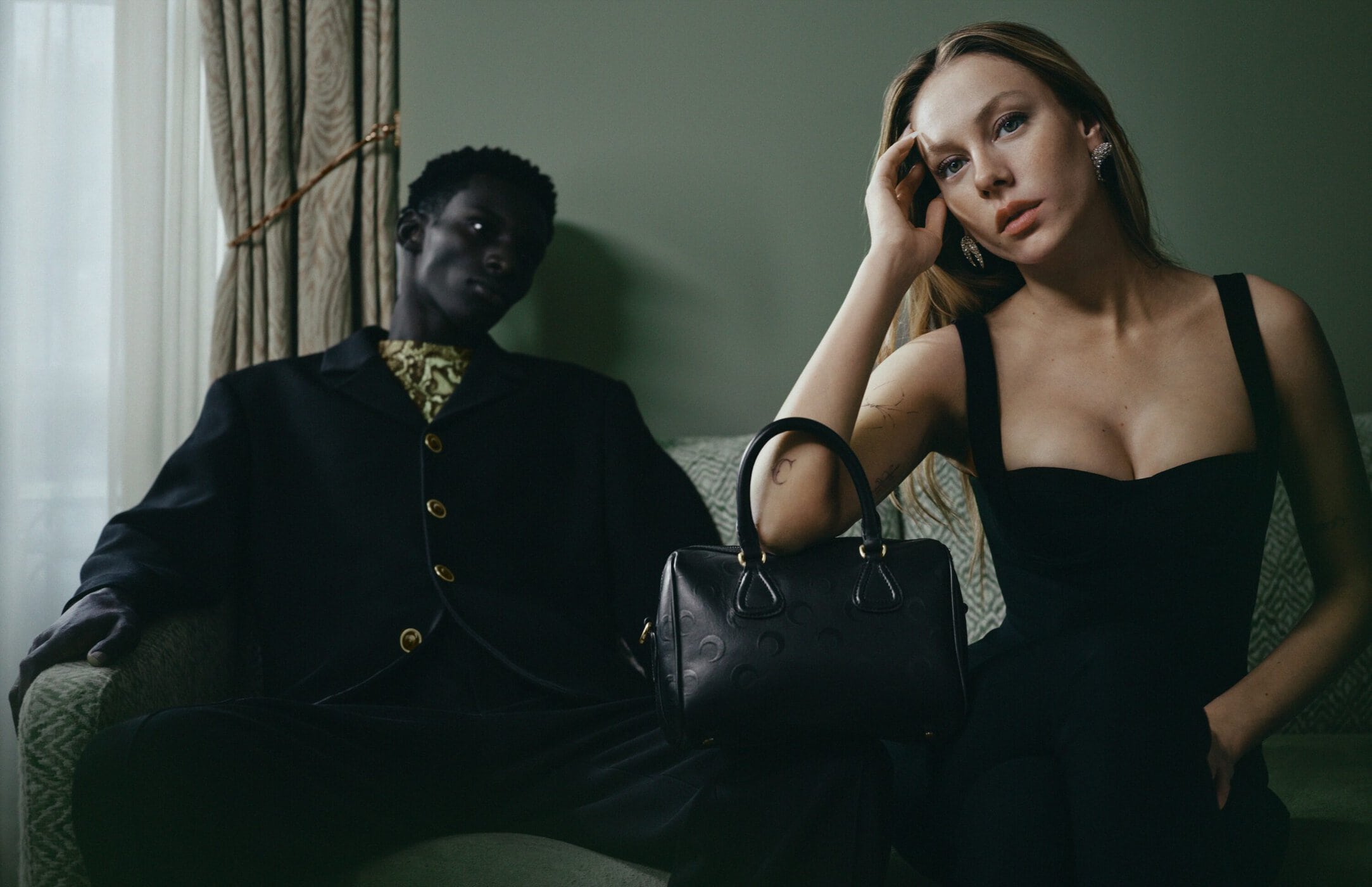 A woman in a black dress resting her arm on a black handbag, with a man seated in a dark jacket in the background.