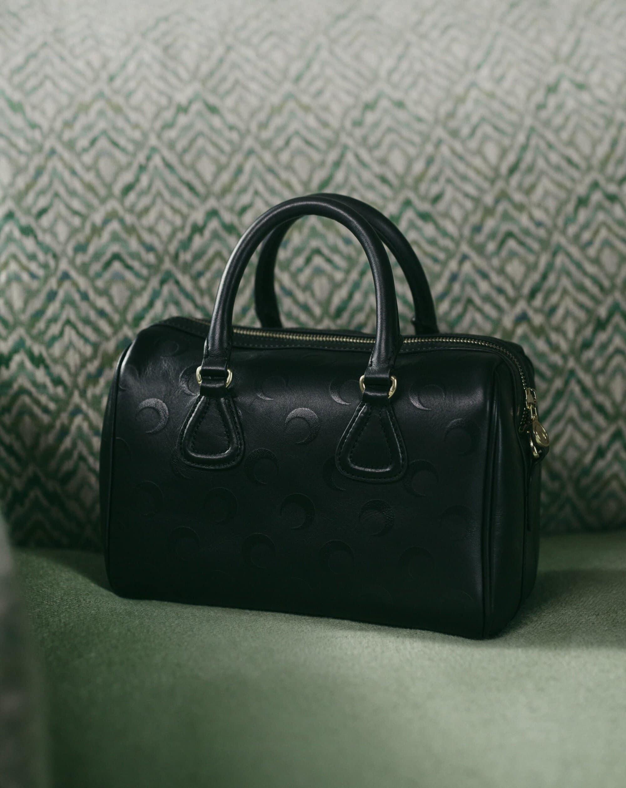 Black leather handbag with crescent moon debossed pattern resting on a green upholstered surface.