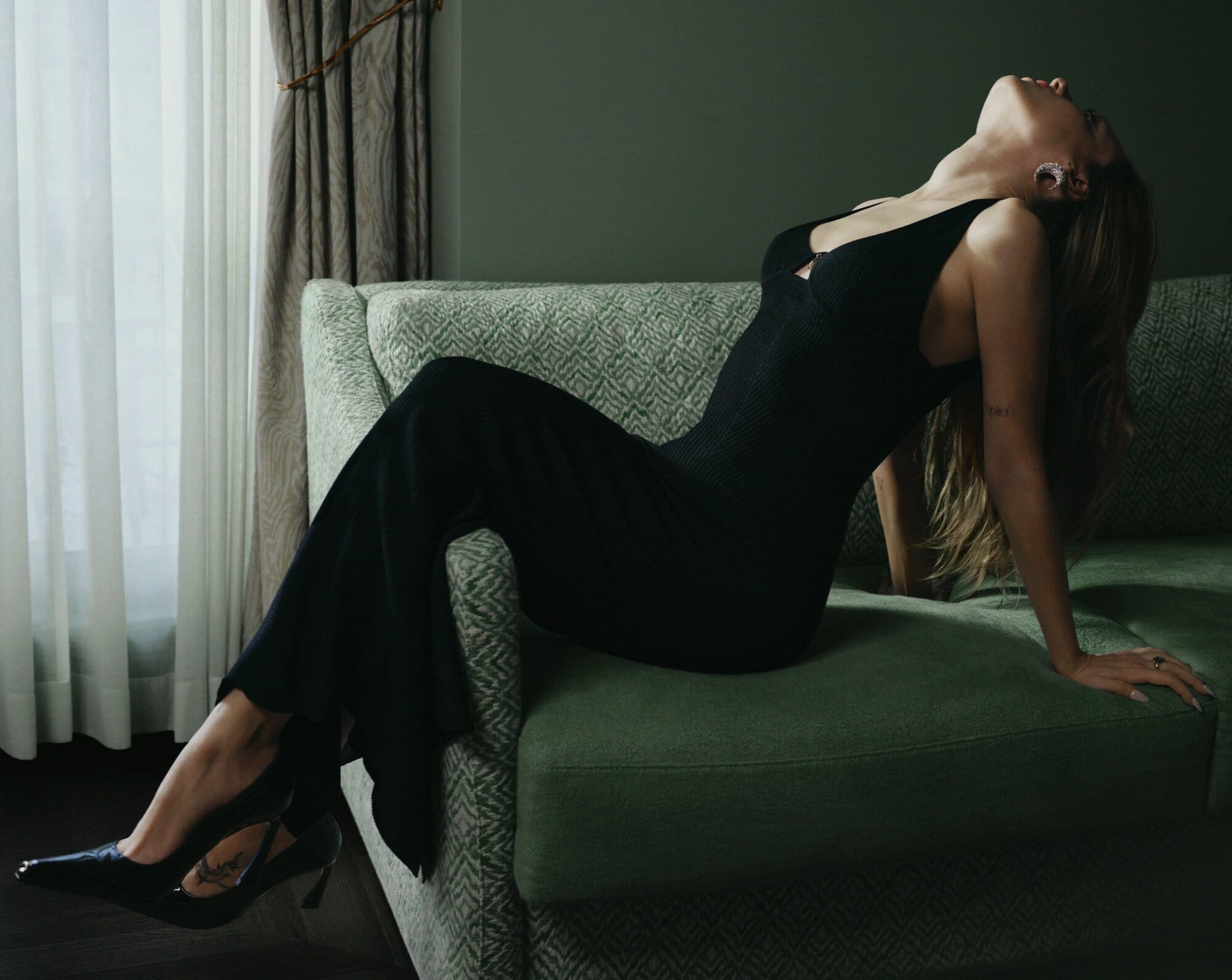 Model in a black dress reclining on a patterned green sofa in an interior setting near a window with sheer curtains.