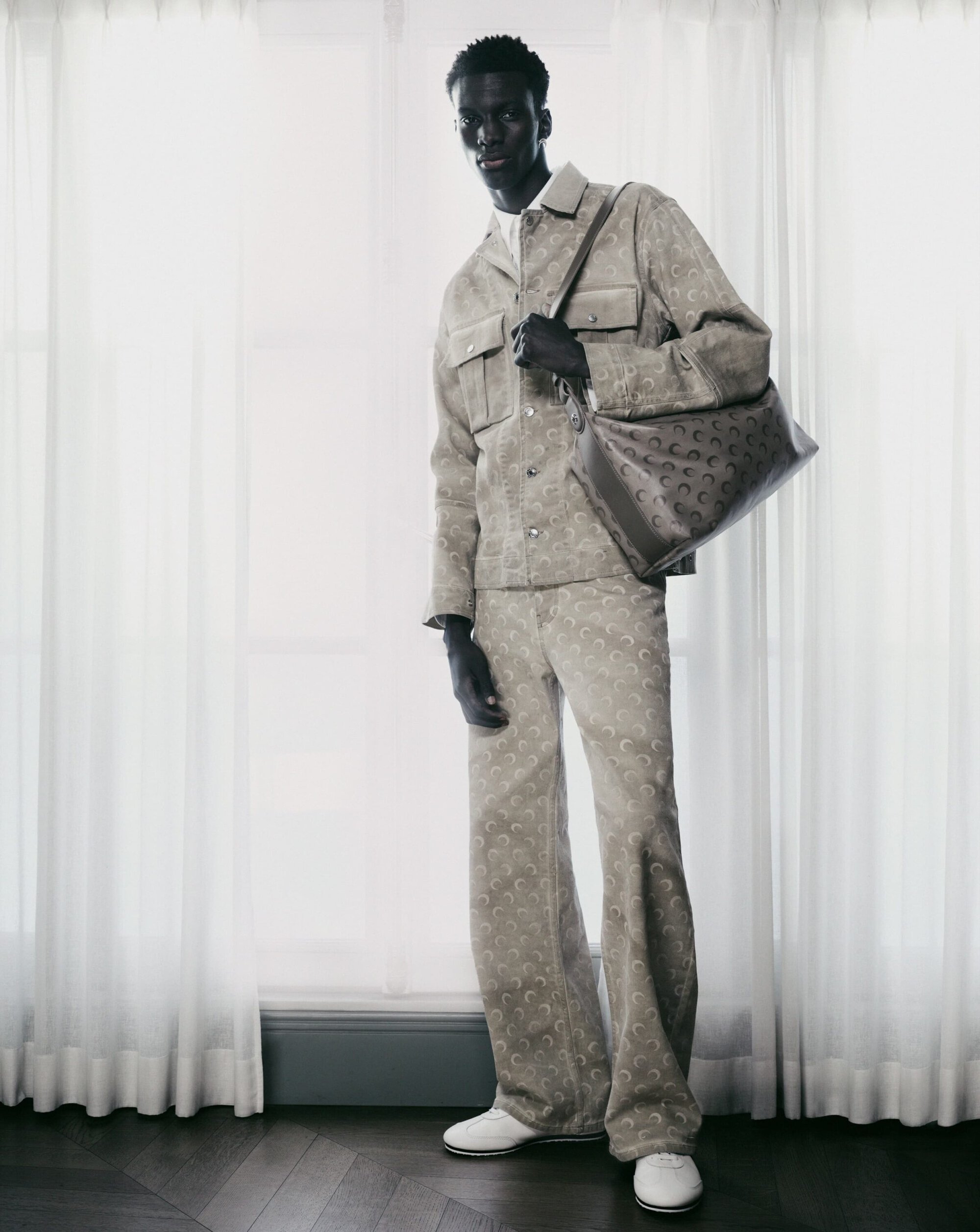 Model Momo Ndiaye in matching printed jacket and trousers holding a shoulder bag against white curtains