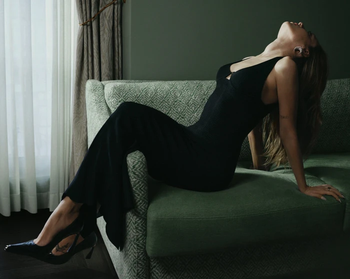 Model in a black dress reclining on a patterned green sofa in an interior setting near a window with sheer curtains.