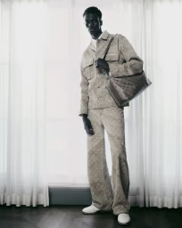 Model Momo Ndiaye in matching printed jacket and trousers holding a shoulder bag against white curtains